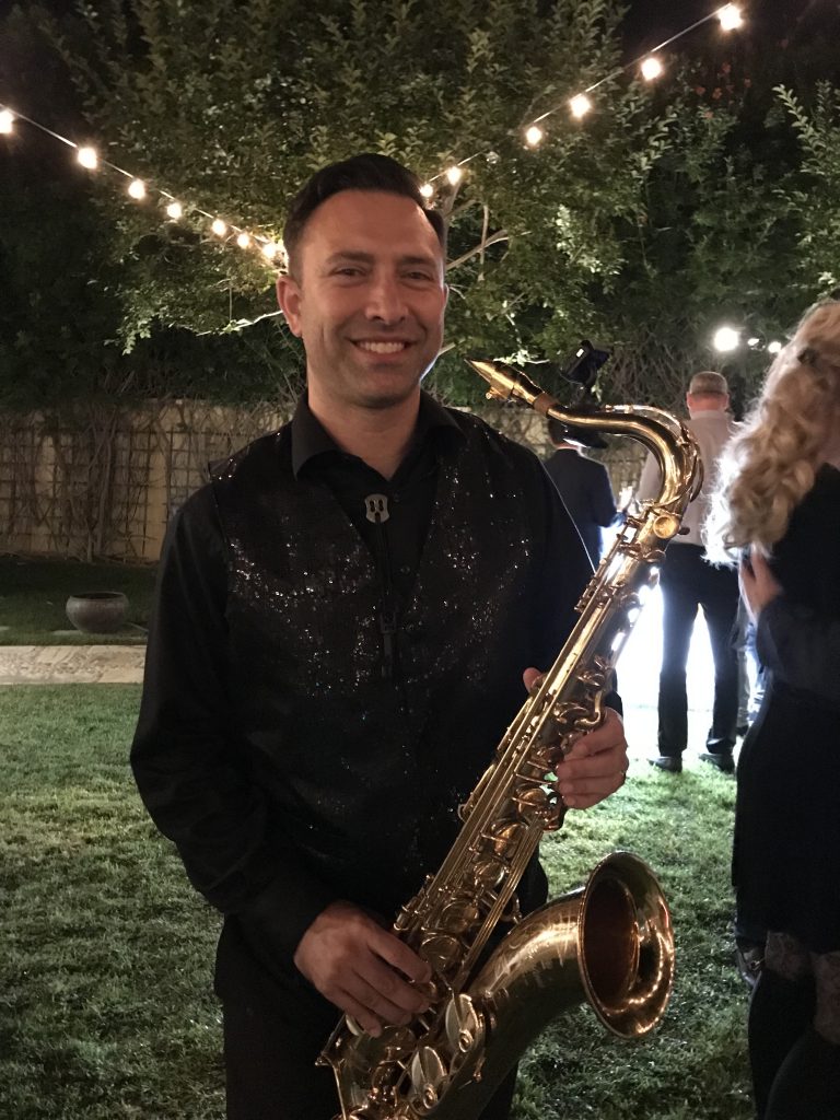 Man smiling with saxophone in backyard with party lighting and guests mingling over cocktails